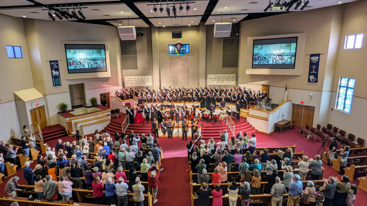 A choir stands on stage after finishing their performance. The audience is visible in the foreground, giving a standing ovation, with many people applauding enthusiastically. The atmosphere is one of excitement and appreciation for the performance.